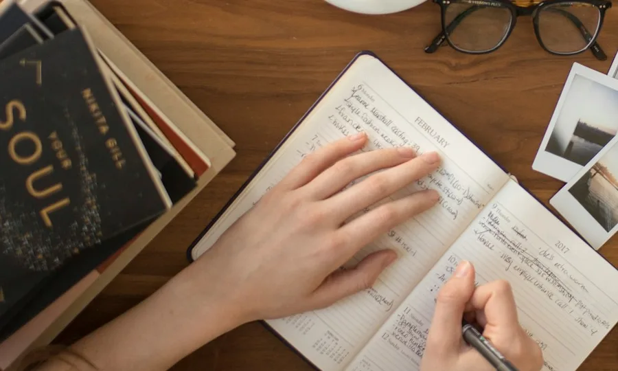 Unas manos tomando notas en un cuaderno, junto a una pila de libros sobre un escritorio de madera